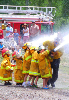 Children dressed as firefighters using a hose, with onlookers and a real fire truck in the background.