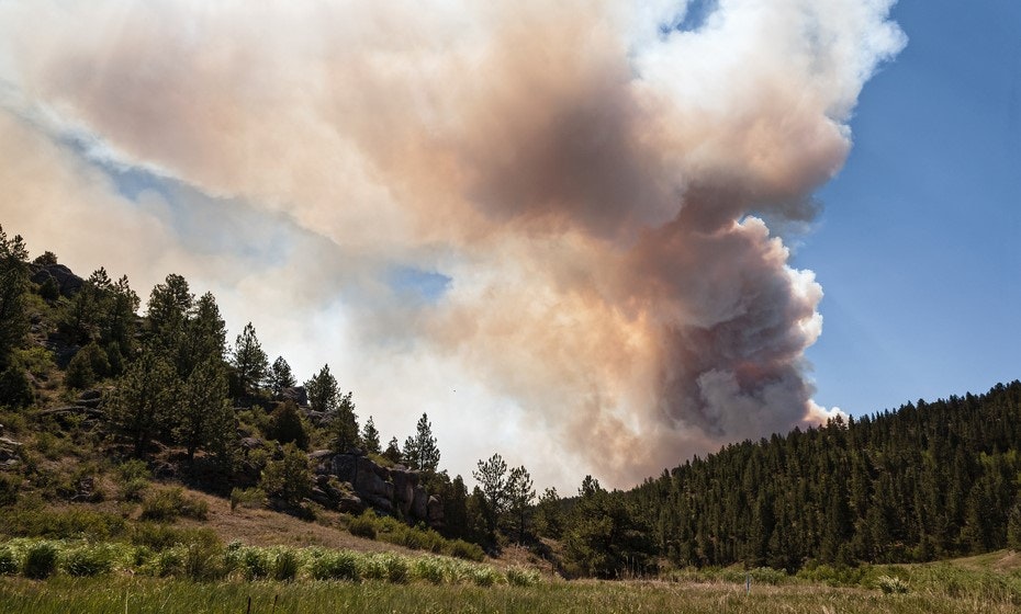 A large wildfire with thick smoke over a forested hillside under a blue sky.