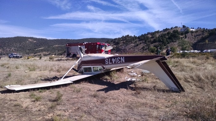 Crashed small aircraft in a field with emergency vehicle in the background.