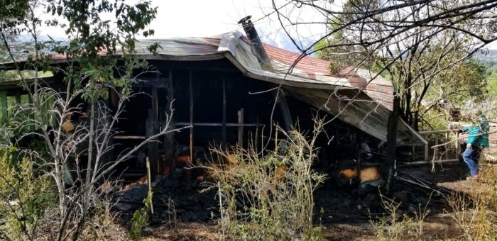 A damaged house with a collapsed roof and charred remains, indicating a recent fire.
