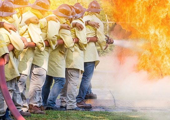 Firefighters in training combat flames with a hose.