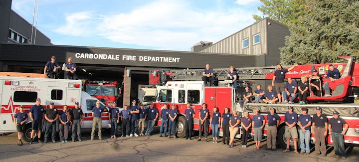 Group of firefighters posing with fire trucks and ambulance in front of a fire department building.