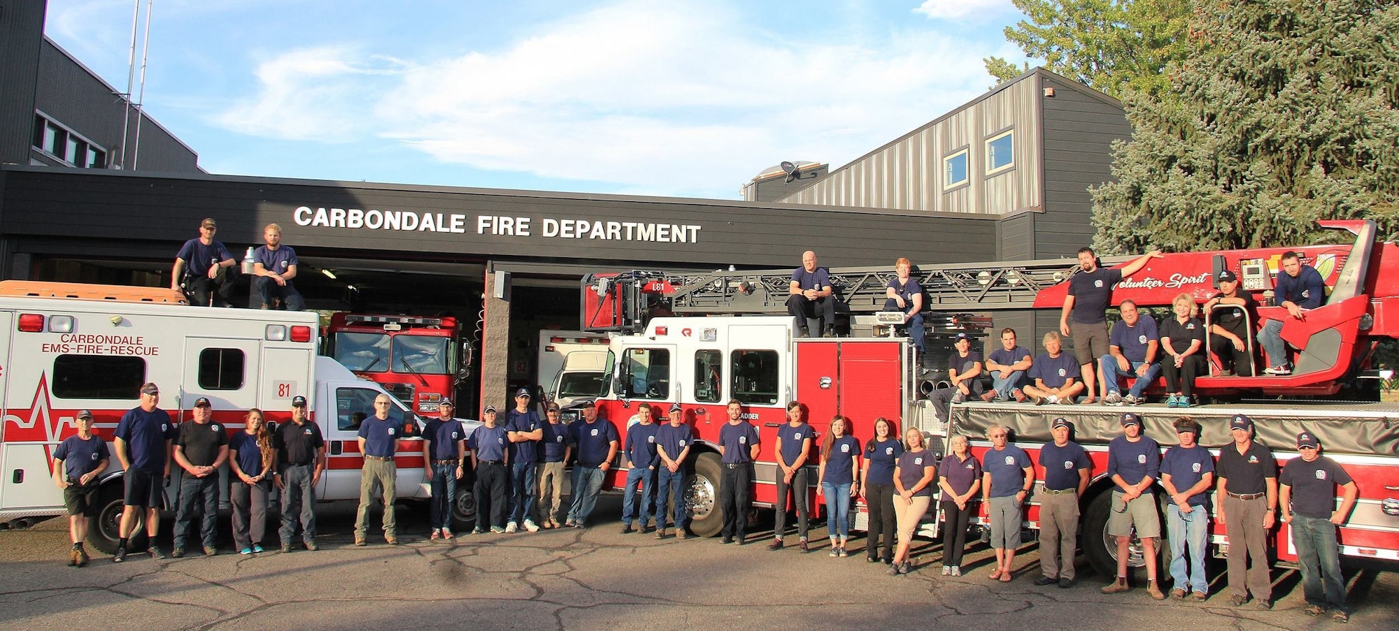 Group of firefighters posing with fire trucks and ambulance in front of a fire department building.