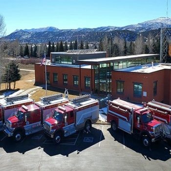 A fire station with three fire trucks parked outside, against a backdrop of mountains with snowy peaks.