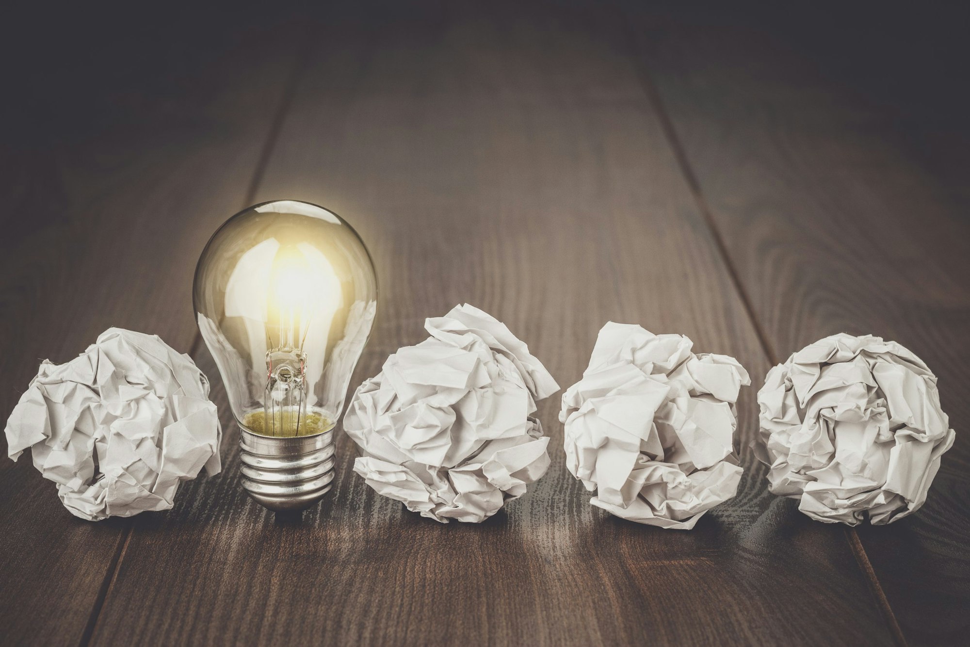 A glowing light bulb stands out among crumpled paper balls on a wooden surface.
