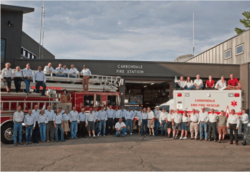 A group of firefighters in front of their station with a fire truck and an ambulance.