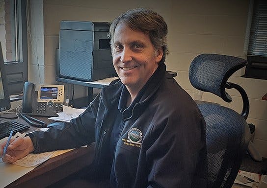 A smiling person at a desk with papers, wearing a black jacket with an emblem, next to a phone and computer.