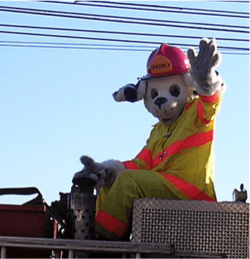 Person in a firefighter-themed mascot costume waving from a vehicle.
