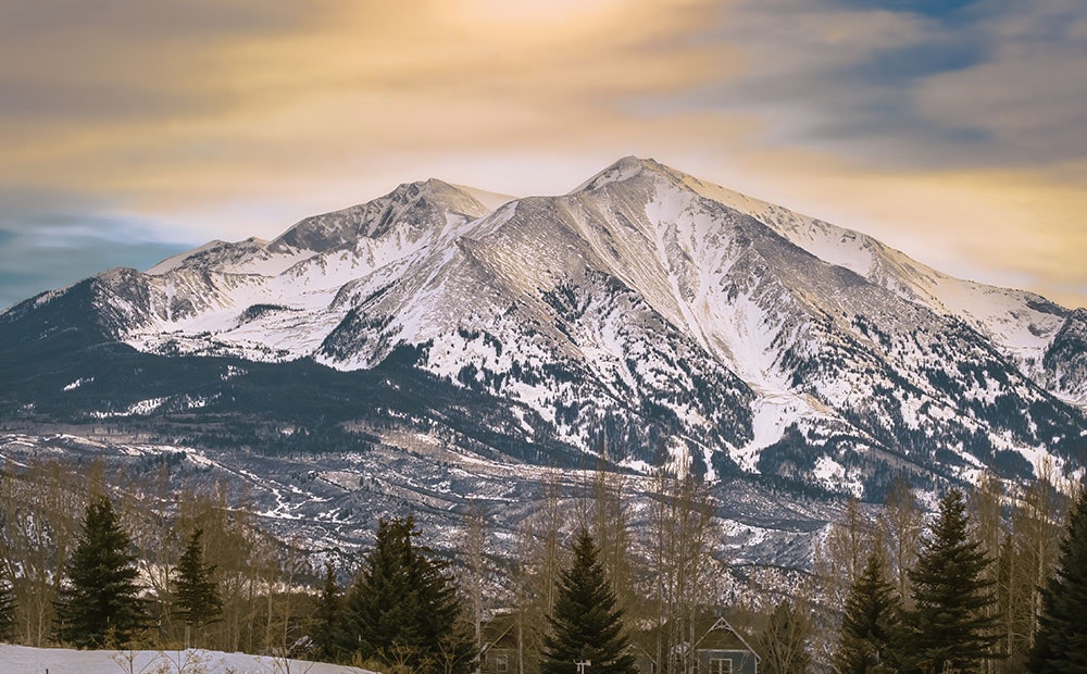 Snow-covered mountains with a golden sky and forest in the foreground.