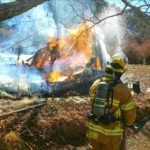 A firefighter watching a large fire consuming a structure amid trees.