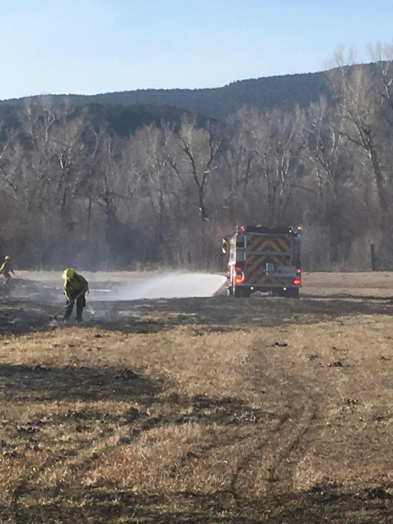 A firefighter hoses down a field with a fire truck in the background, set against a backdrop of trees and hills.