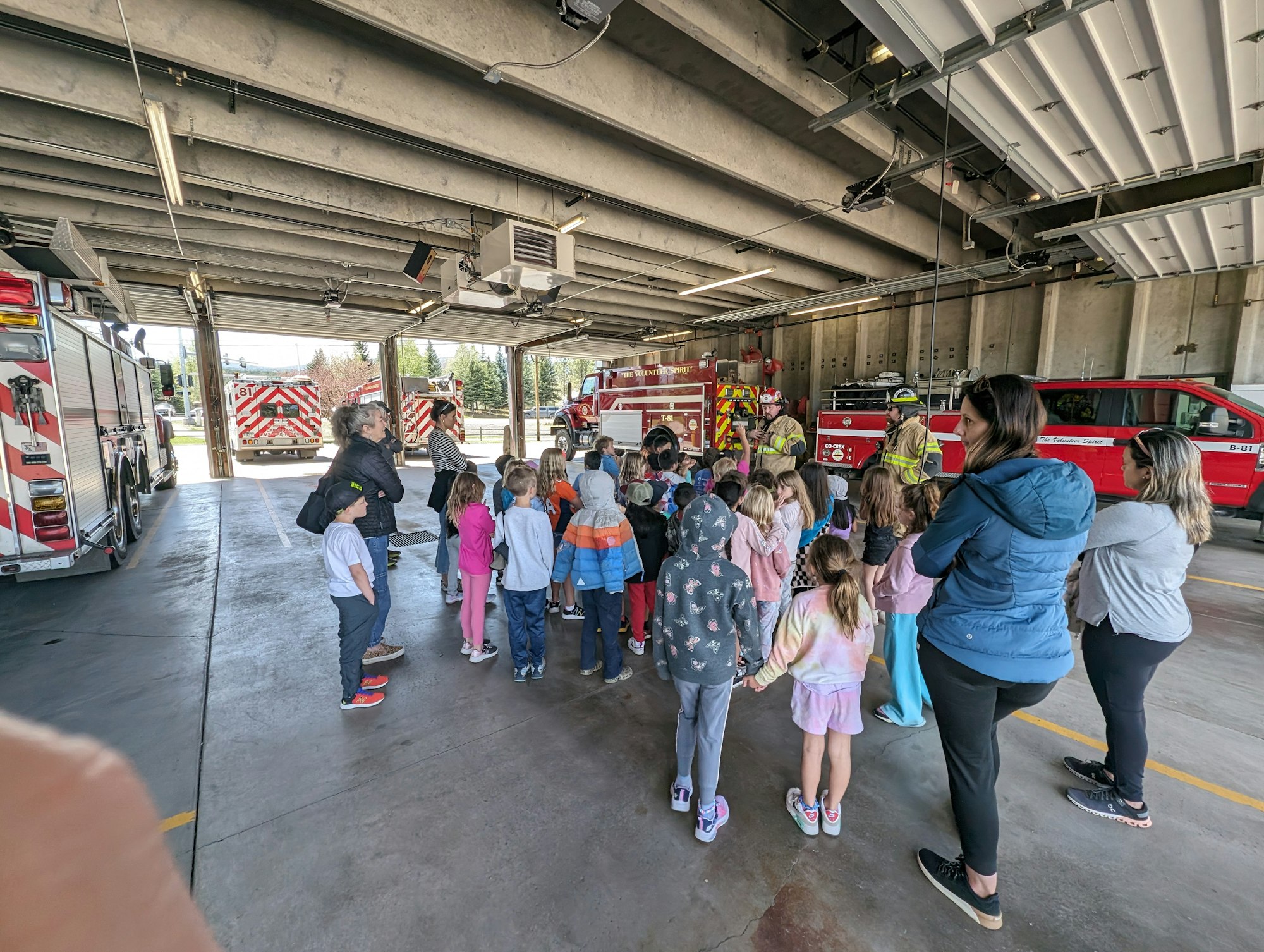 Children and adults gather inside a fire station with several fire trucks, two firefighters addressing the group.