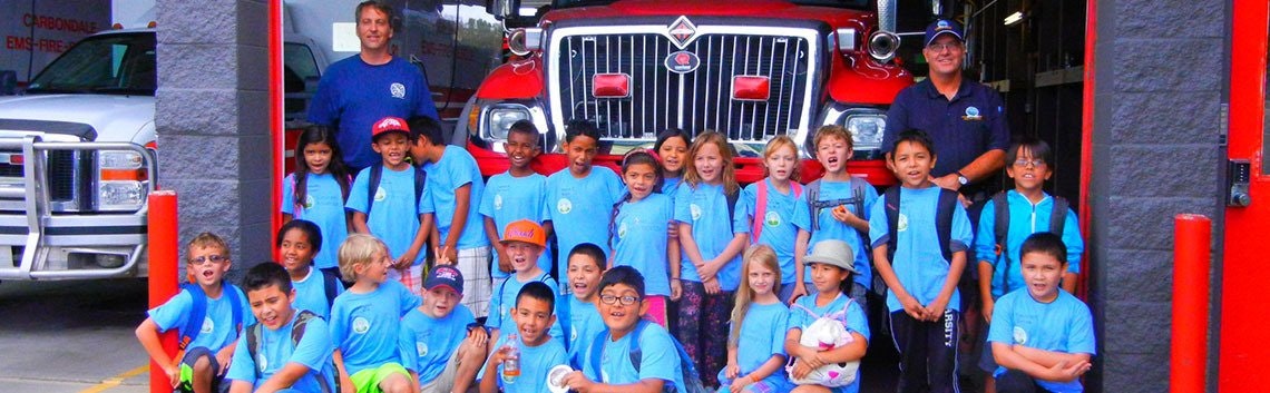 A group of children and firefighters in front of a fire truck at a fire station.