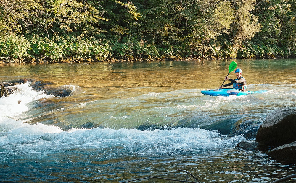 Person kayaking on a river with mild rapids, surrounded by greenery.