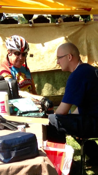 Two people sitting, one in cycling gear, conversing at a table with personal items and snacks.