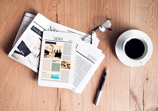 A newspaper, eyeglasses, a cup of coffee, and a pen on a wooden surface.