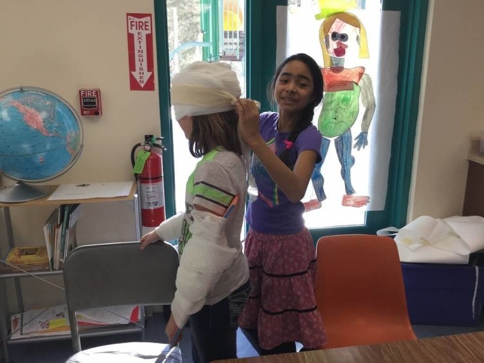 Two kids in a classroom, one wrapping a bandage around the other's head, with artwork and globe in the background.