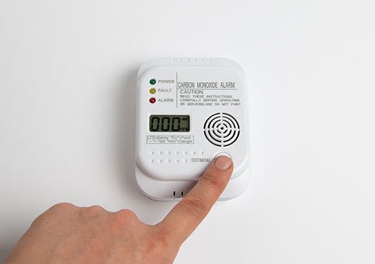 A finger is pressing the test button on a wall-mounted carbon monoxide alarm showing a clear digital display.