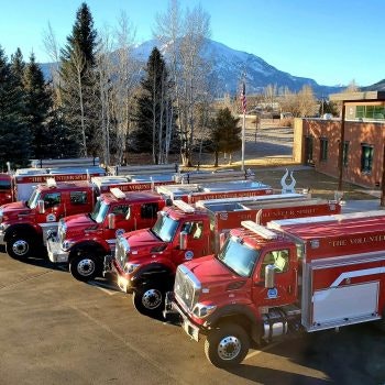 A lineup of red fire trucks in front of a building with mountains in the background.