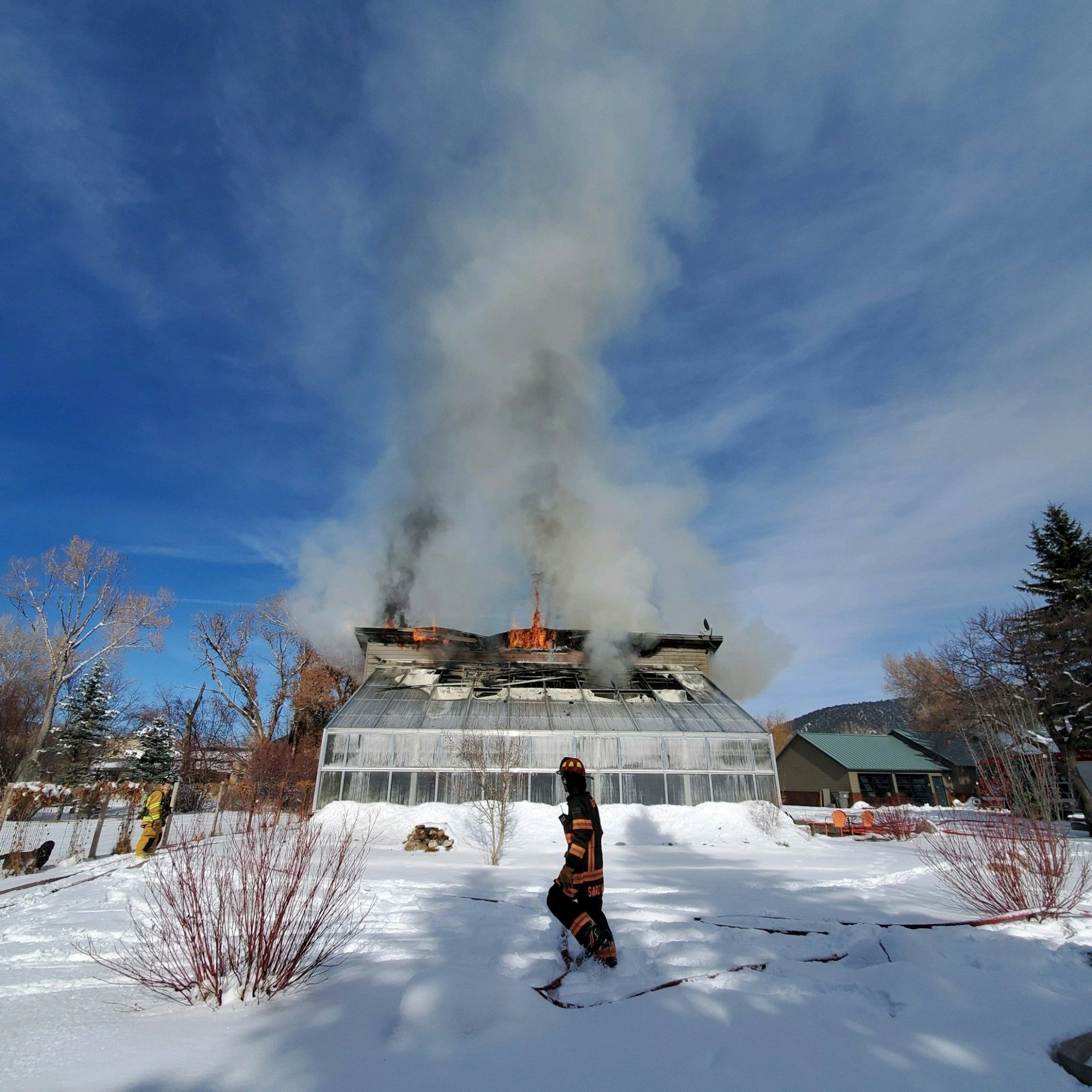 A firefighter observes a building with a smoking roof engulfed in flames, amidst a snowy landscape under a clear blue sky.