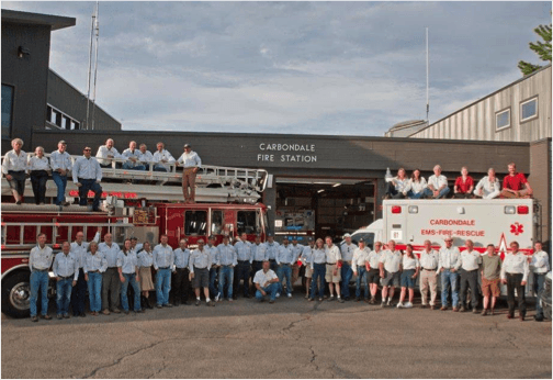 Group of firefighters posing in front of their station with fire trucks and an ambulance.