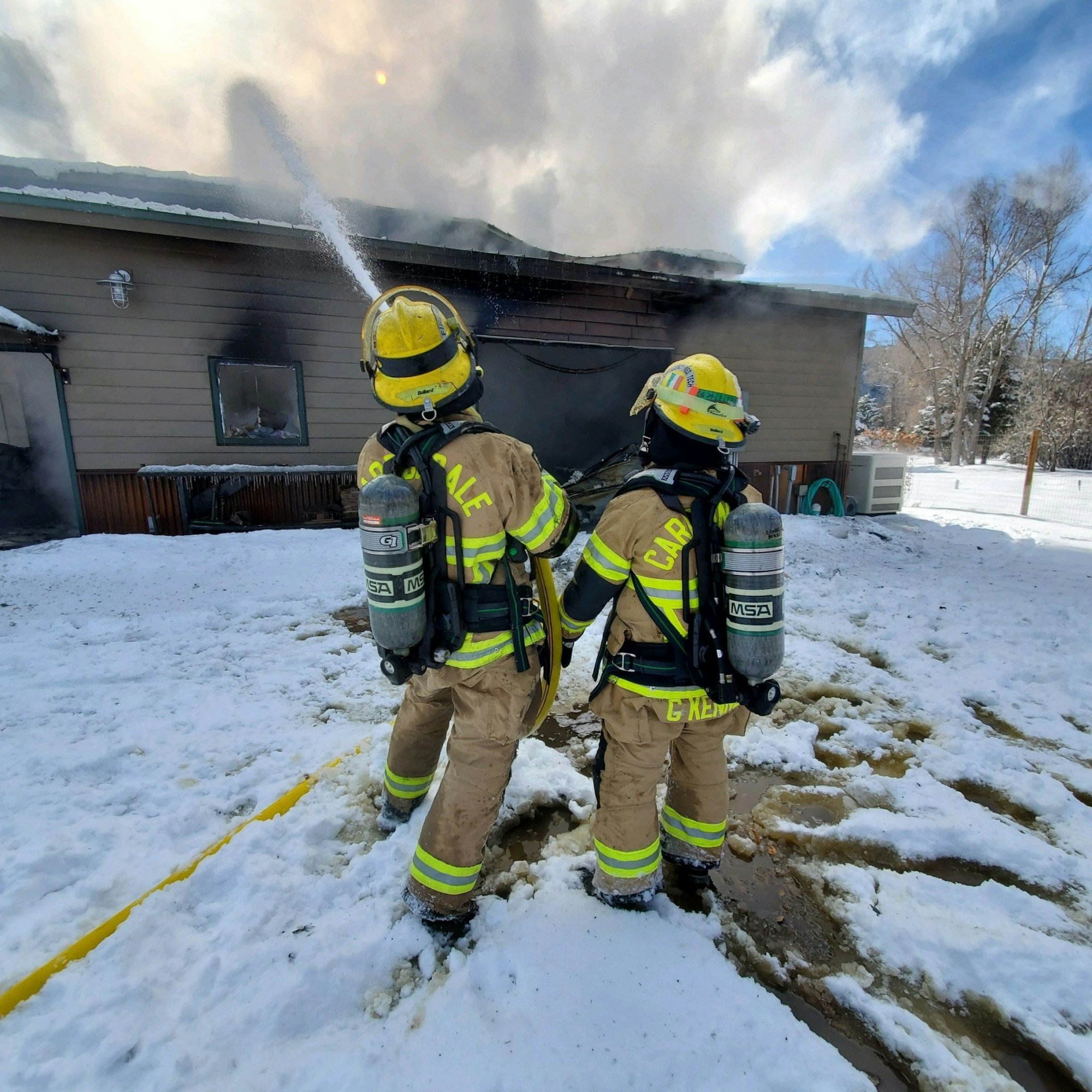 Two firefighters with a hose confronting a house fire, surrounded by snow.