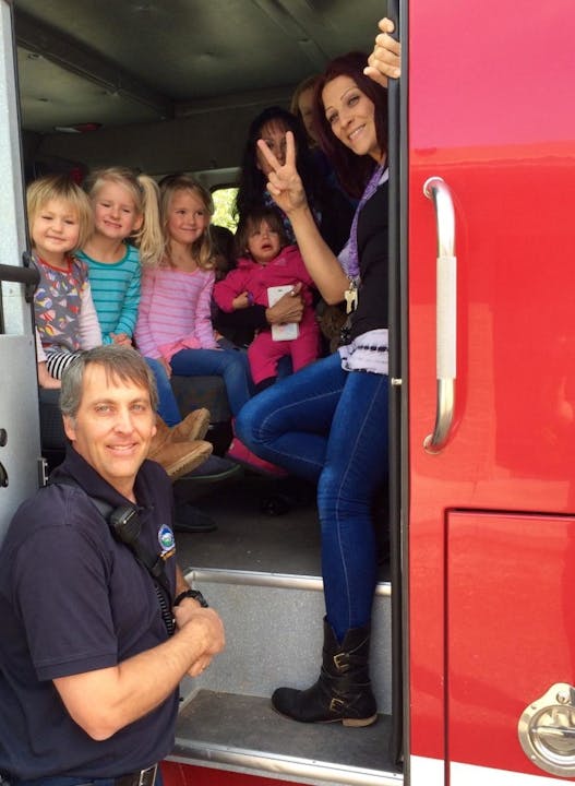 A group of kids with adults inside a fire truck, with a firefighter standing by the door.