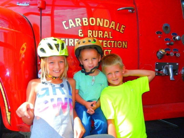 Three children smiling in front of a red fire truck labeled "Carbondale & Rural Fire Protection."