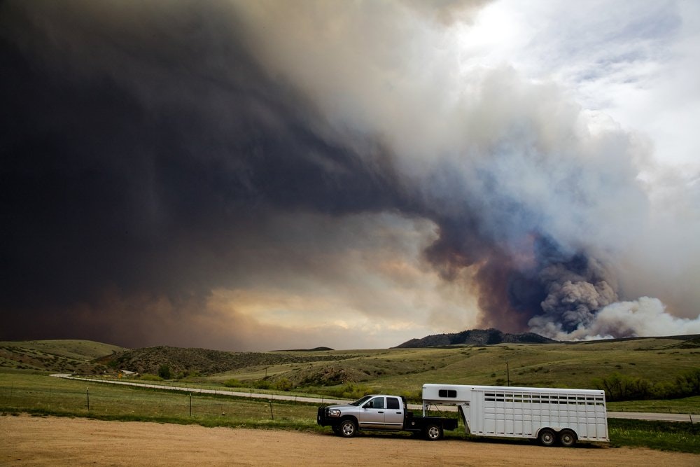 A truck with a trailer parked in a field under a dark, ominous sky with a large smoke plume from a wildfire in the distance.