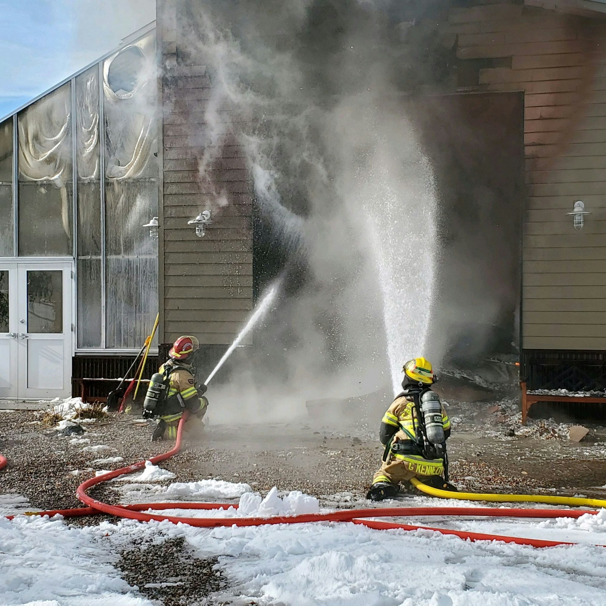 Firefighters with hoses extinguishing a building fire, surrounded by snow, steam, and smoke.