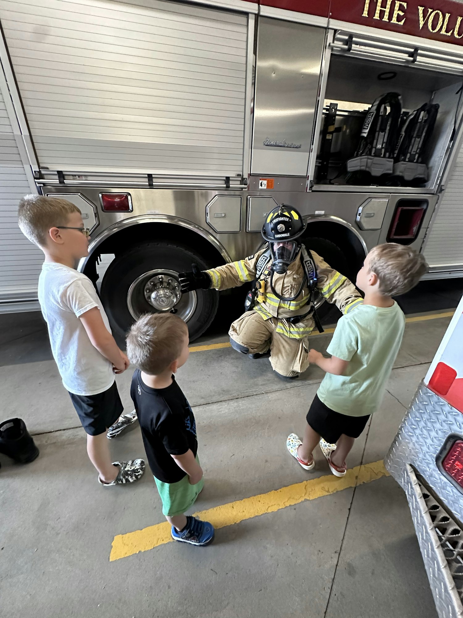 A firefighter in gear talks to three children in front of a fire truck.