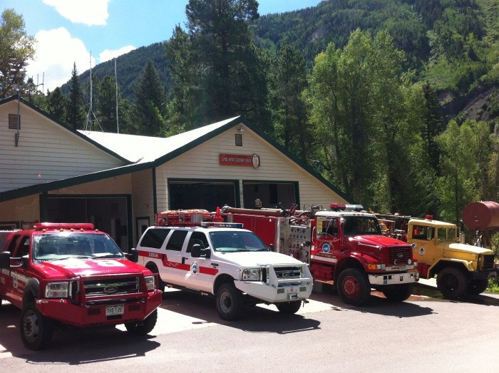 Fire station with emergency vehicles and trucks parked outside, surrounded by trees and mountains.