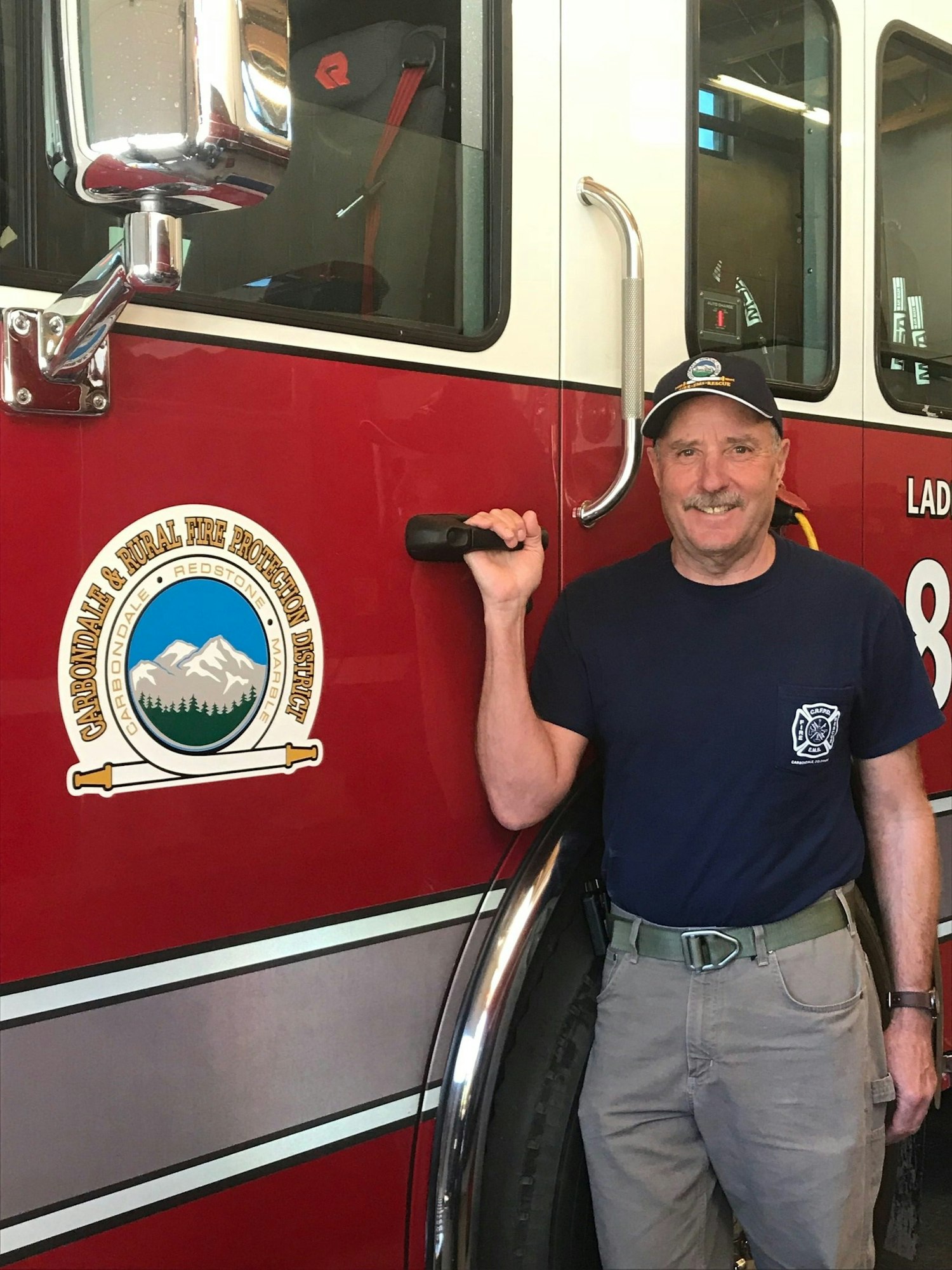 Man with a cap standing next to a fire truck, holding a walkie-talkie.