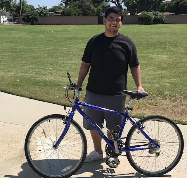 A person stands on a sidewalk holding a blue bike, with a grassy field in the background.