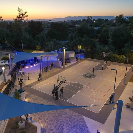 Basketball court with people playing, surrounded by trees and lit up at dusk, with a nearby gathering area under blue lights.