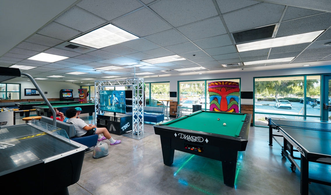 Game room with pool table, ping pong, air hockey, TV, and people seated. Bright, modern decor with colorful wall art and large windows.