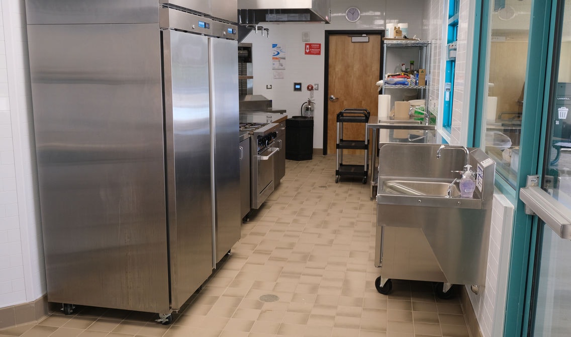 A commercial kitchen with stainless steel appliances, sink, shelves, cart, and a door at the end.