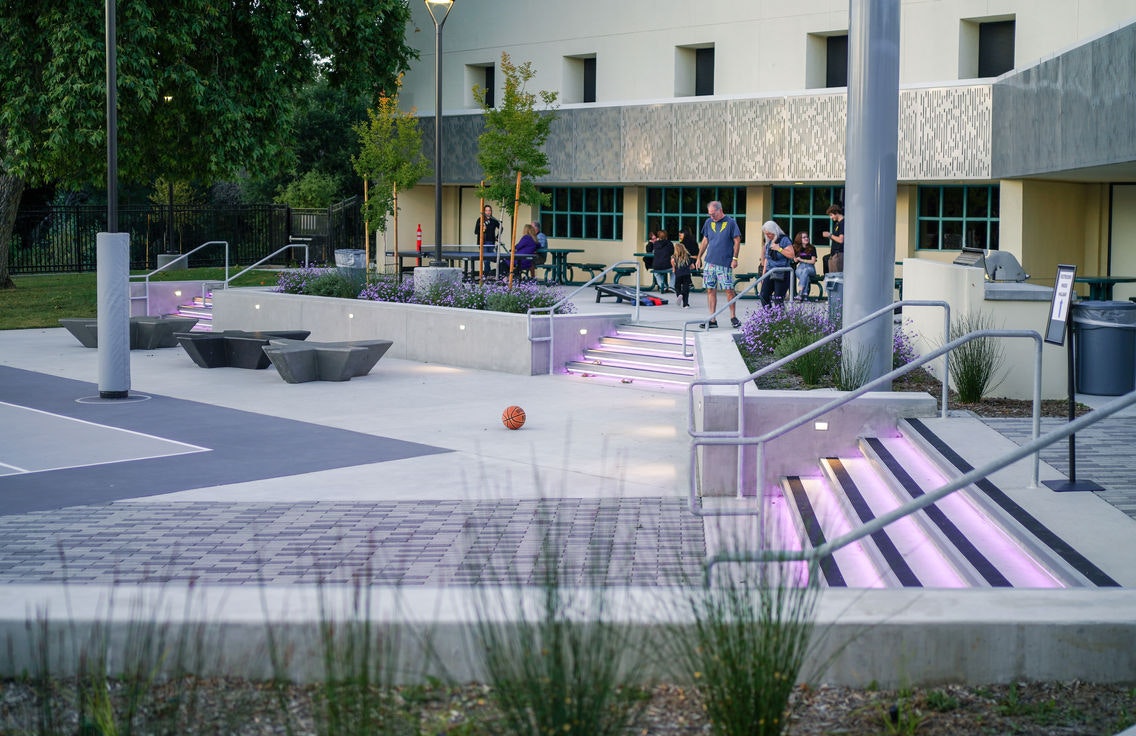 Outdoor space with modern seating, stairs with purple lights, basketball on the ground, and people socializing.