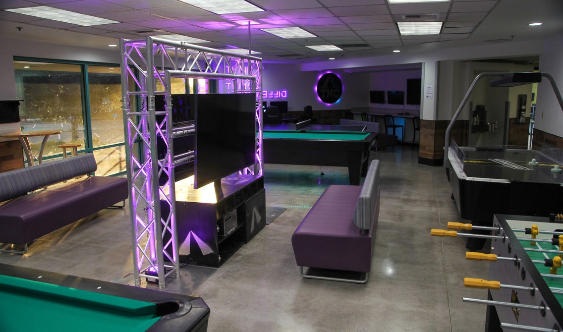 A modern game room with pool tables, a foosball table, TV, sofas, and atmospheric purple lighting.