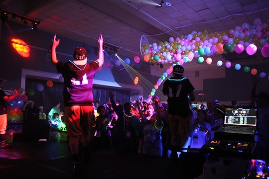 Party scene with people dancing, wearing jerseys, colorful balloons overhead, DJ playing music, and vibrant lighting effects.