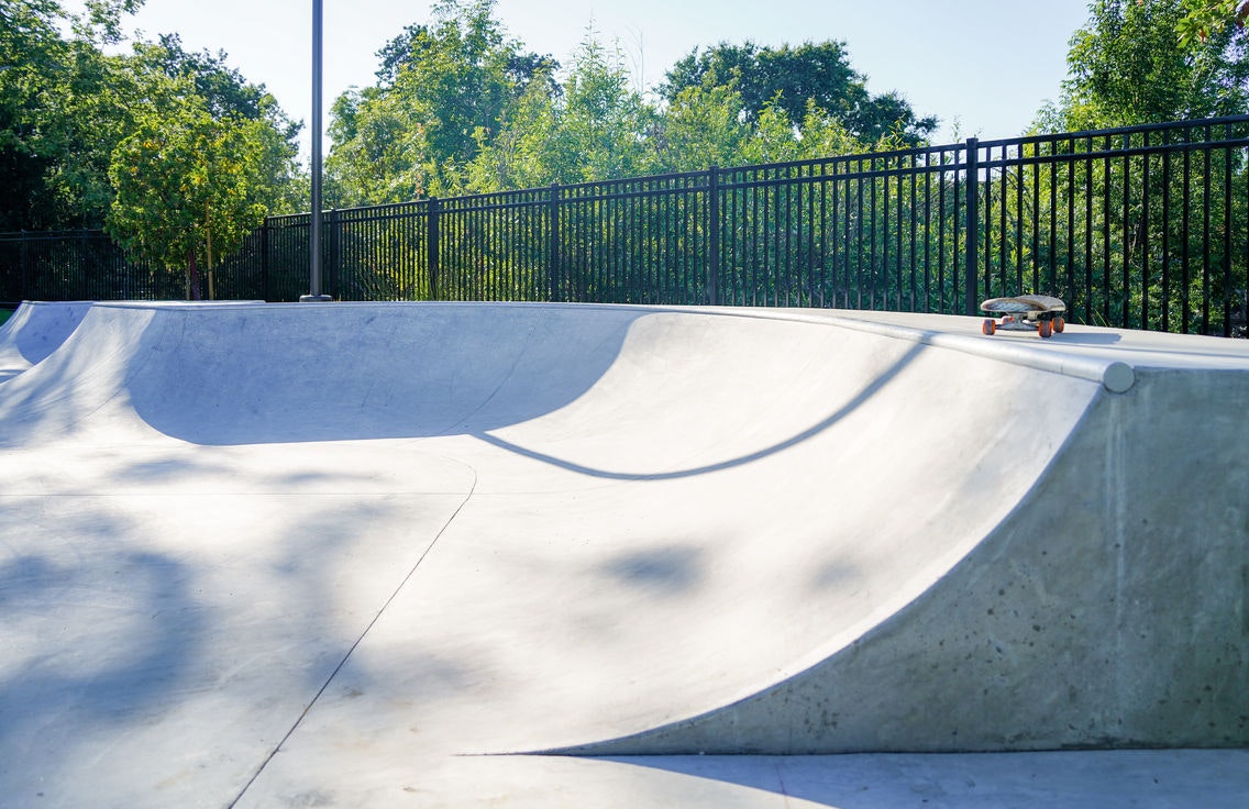 A skateboard on the edge of a concrete skatepark ramp, surrounded by trees and a fence.