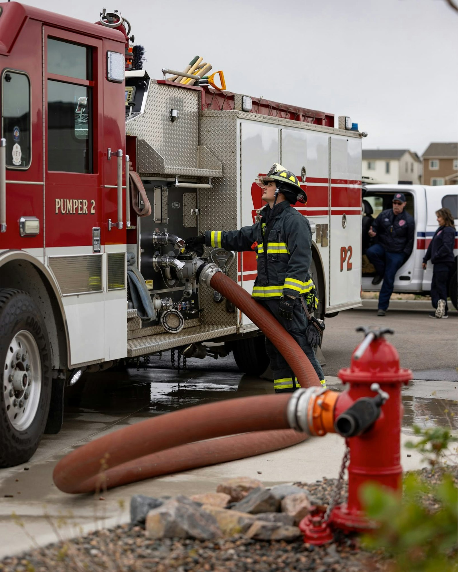 A firefighter operates a hose connected to a fire truck, drawing water from a fire hydrant, with others nearby.