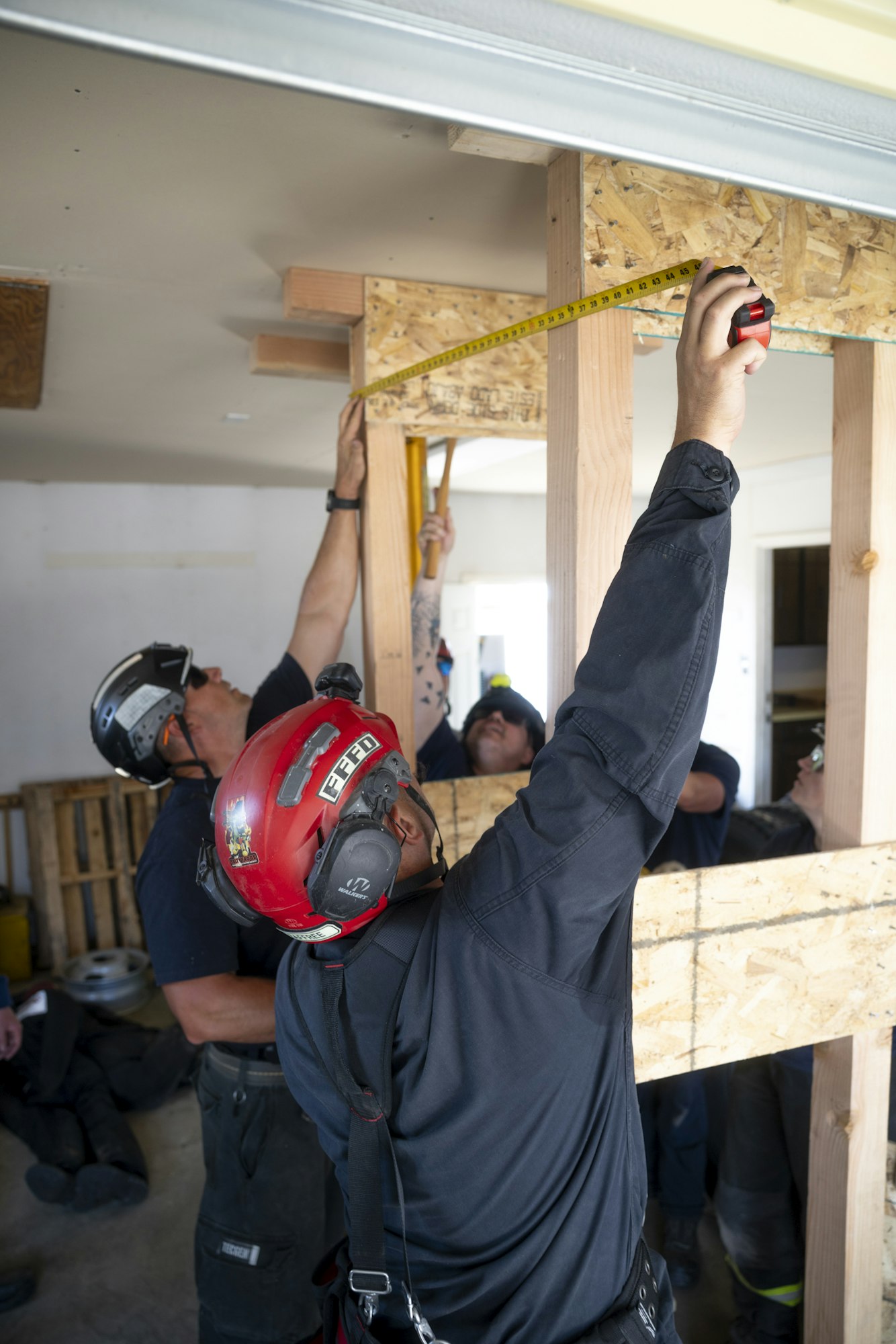 A group of workers measures wooden supports, likely for construction or renovation, while wearing safety helmets and gear.
