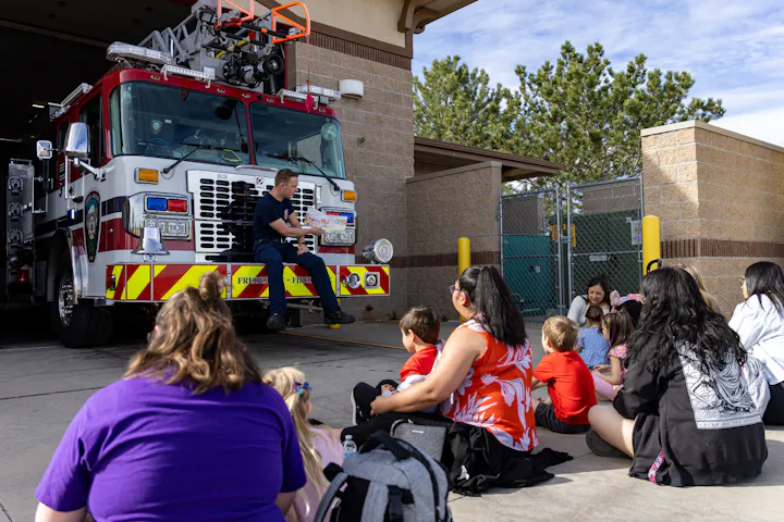 A firefighter engages a group of children and adults in front of a fire truck during an educational event.