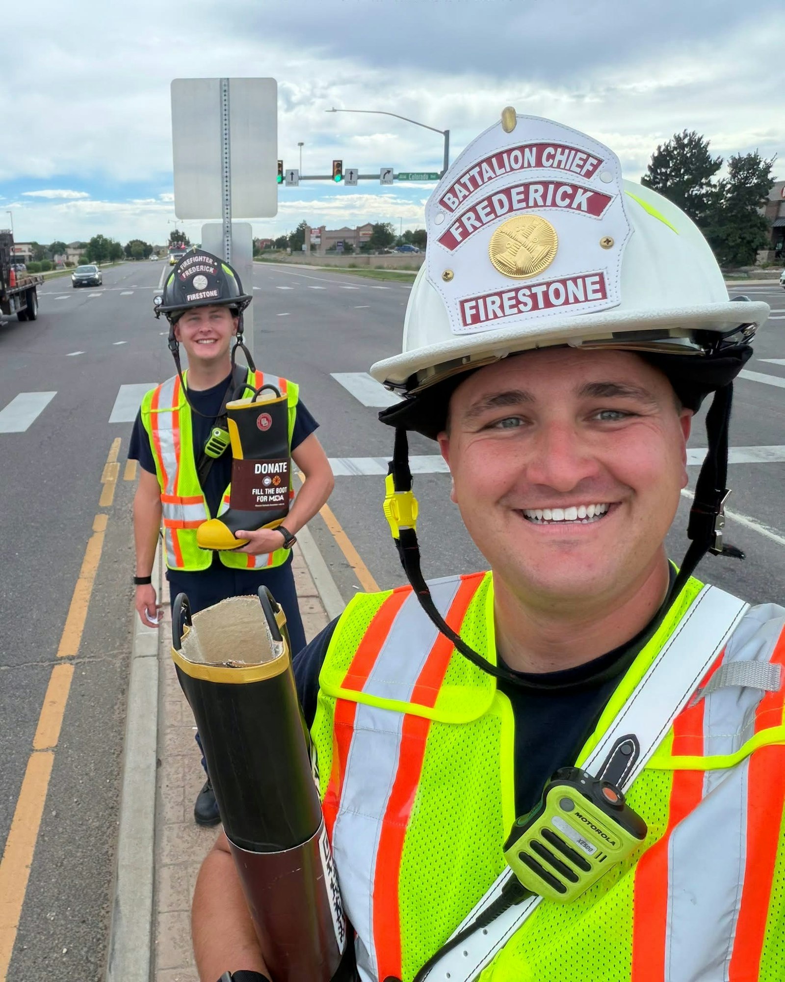 Two firefighters in safety vests smile while fundraising on a roadside, holding a donation container.
