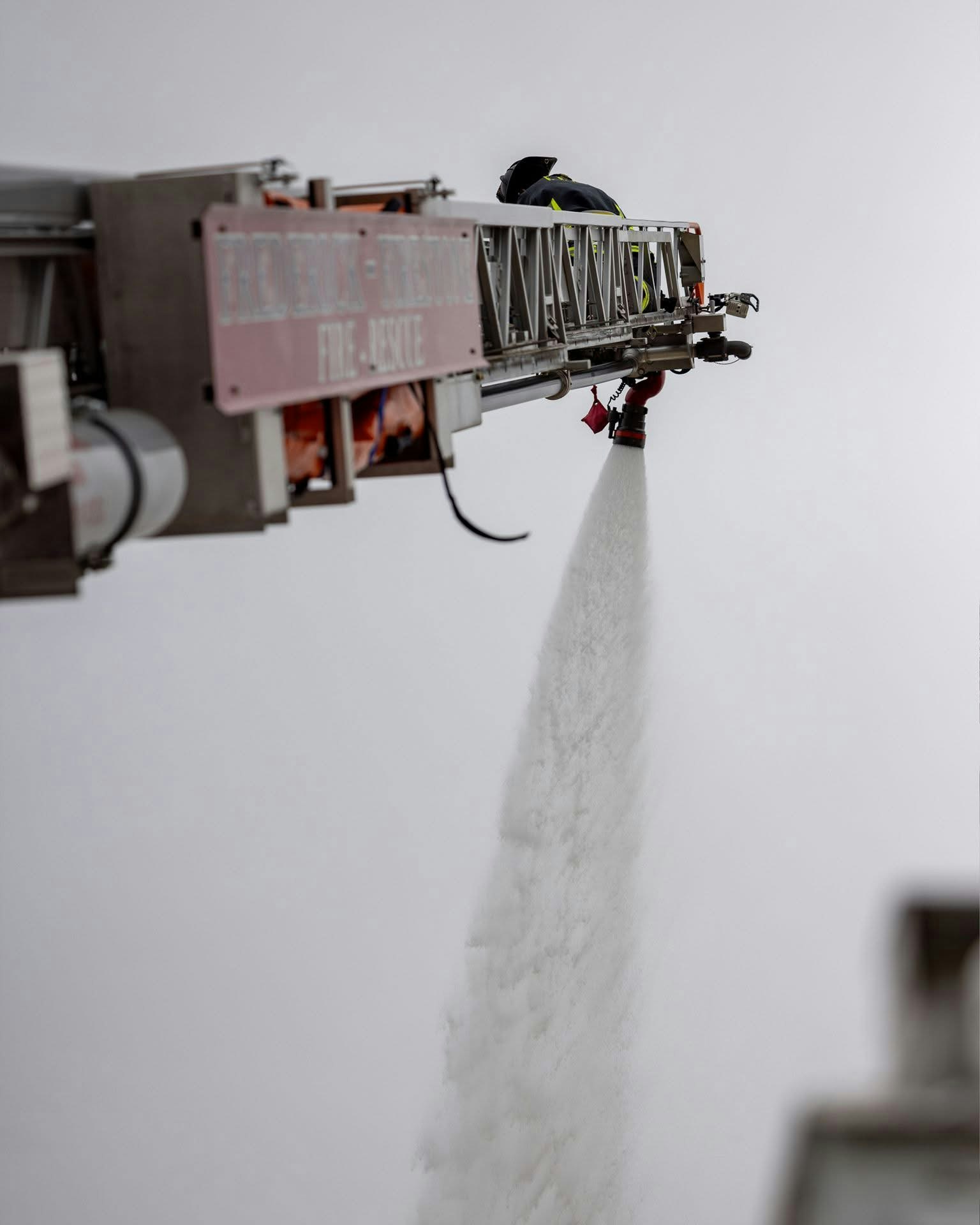 A firefighter operates a ladder truck, spraying water or foam, likely in response to a fire or emergency situation.