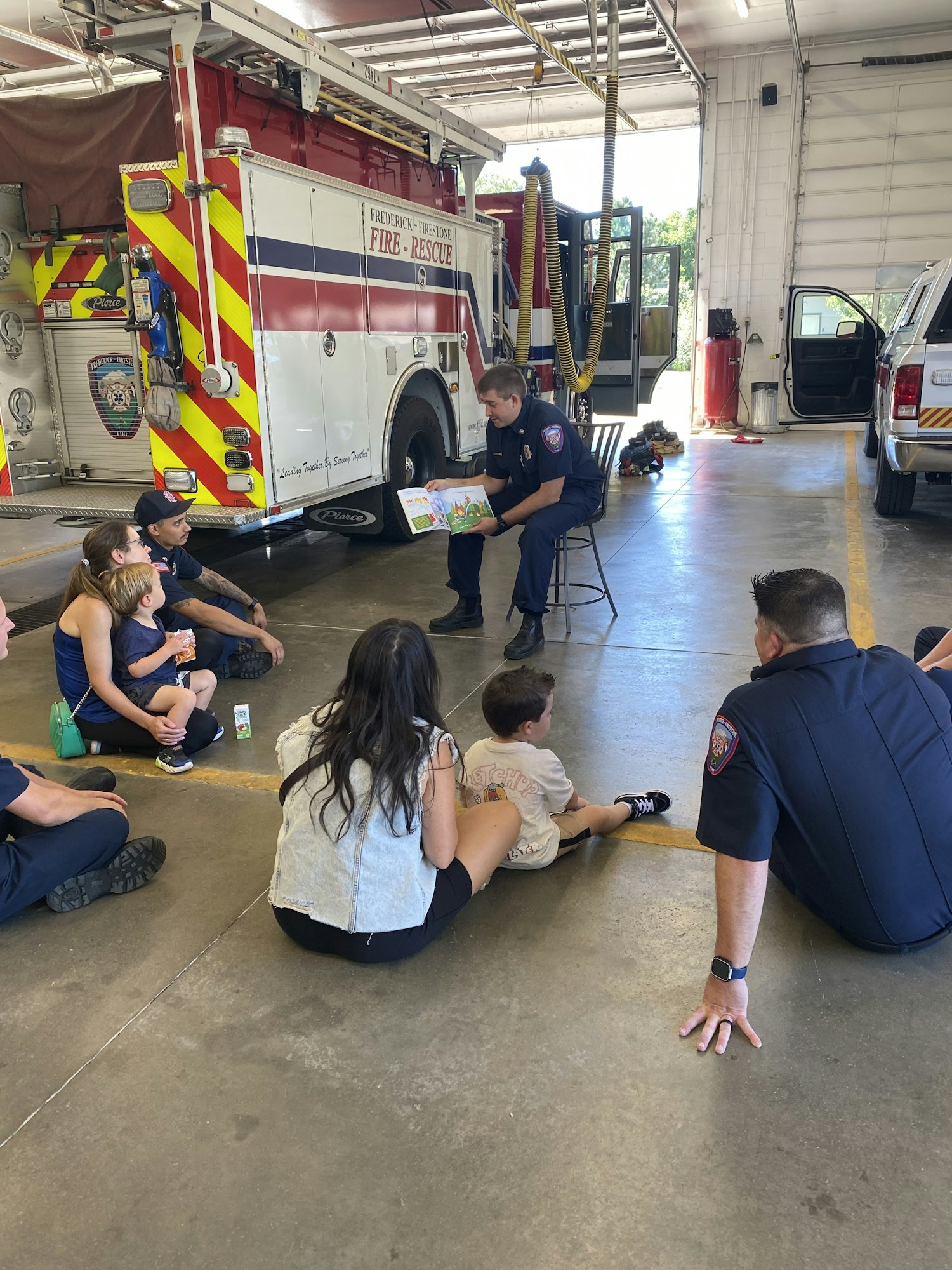 Firefighters sitting with children on a fire station floor, possibly during an educational visit.