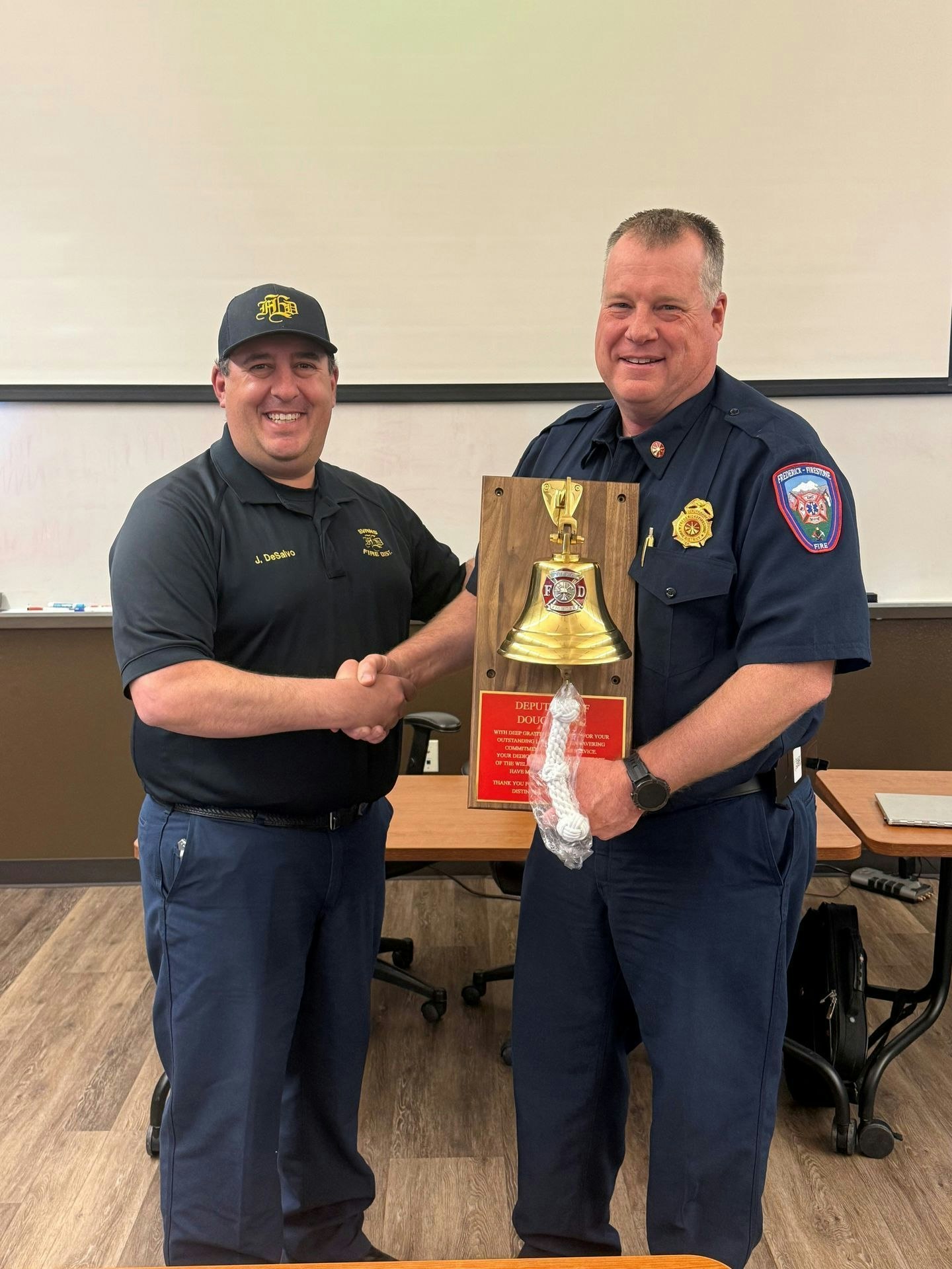 Two men shake hands, one holding an award featuring a bell, in a classroom setting. It celebrates a significant achievement.