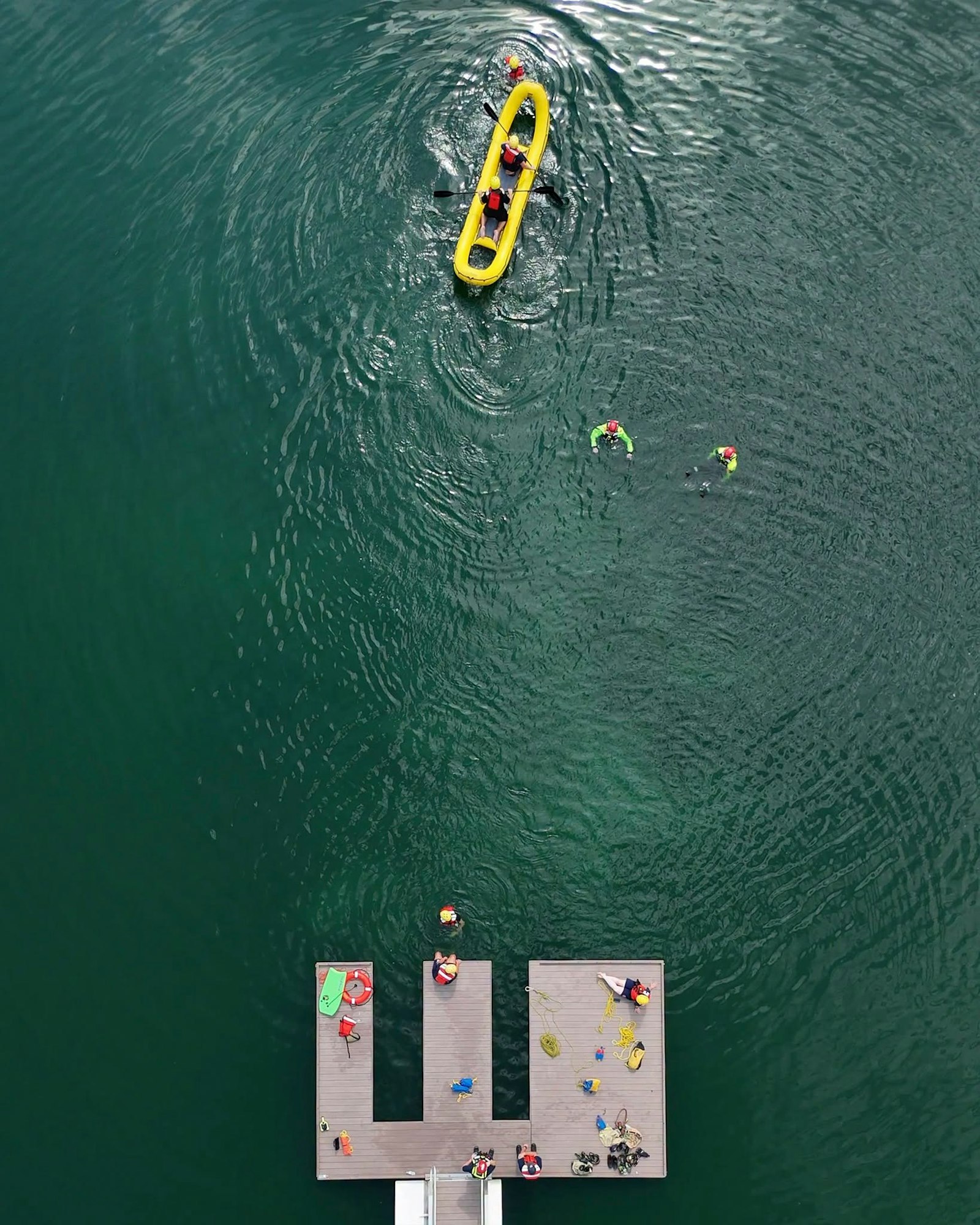 An aerial view shows a dock with people, kayakers, and swimmers in a green lake, creating a lively water scene.