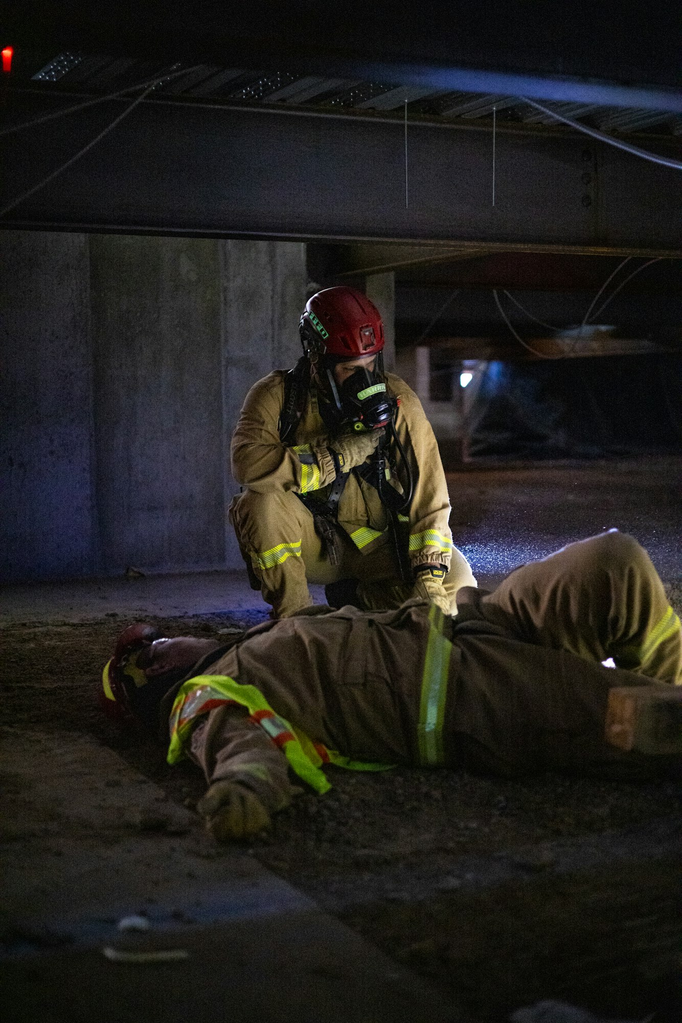 A firefighter, wearing a mask, attends to an unconscious person lying on the ground in a dimly lit area.
